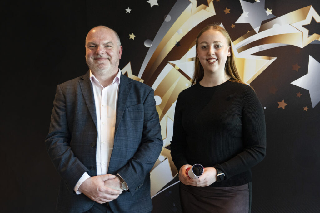 A smiling man in a suit stands next to a tall woman in a black sweater and jeans holding a rolled-up certificate, against a star-patterned background.
