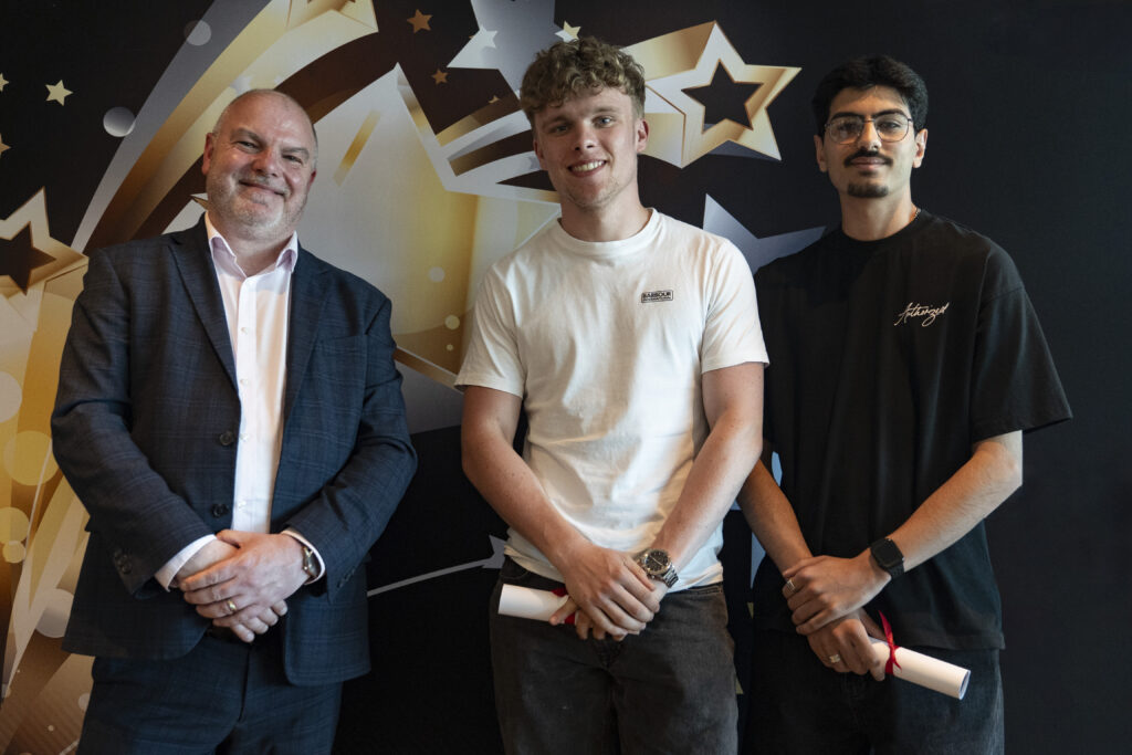  A smiling man in a suit stands between two younger men, both holding rolled-up certificates, against a star-patterned background.