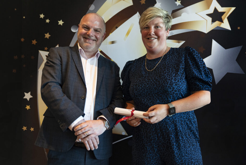  A smiling man in a suit and a smiling woman in a dress holding a certificate, against a star-patterned background.