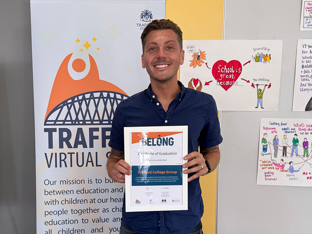 Shaun Deakin holding the Belong Certificate of Graduation in front of a Trafford College Group banner and community posters.