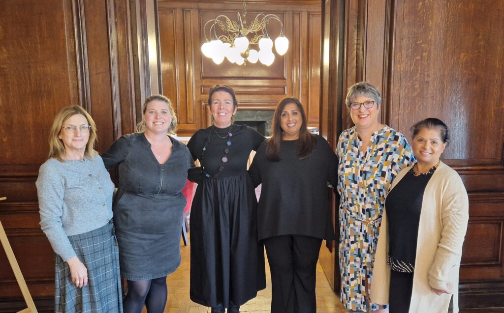 Six people standing side by side in a wood-paneled room with a chandelier, all facing the camera and dressed in professional attire.