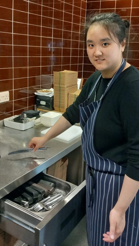Girl wearing an apron in a kitchen, with utensils and equipment visible on the counter.