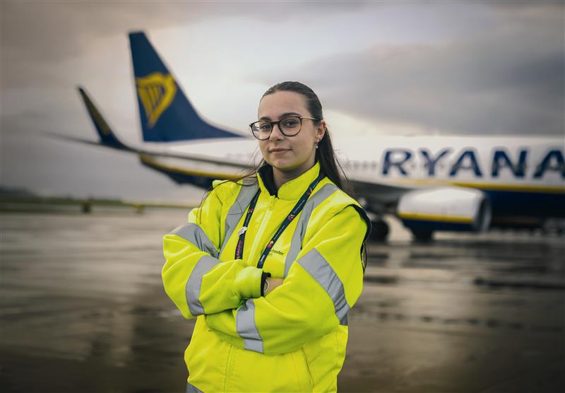 Apprentice in high vis jacket standing in front of a Ryanair plane with her arms folded.