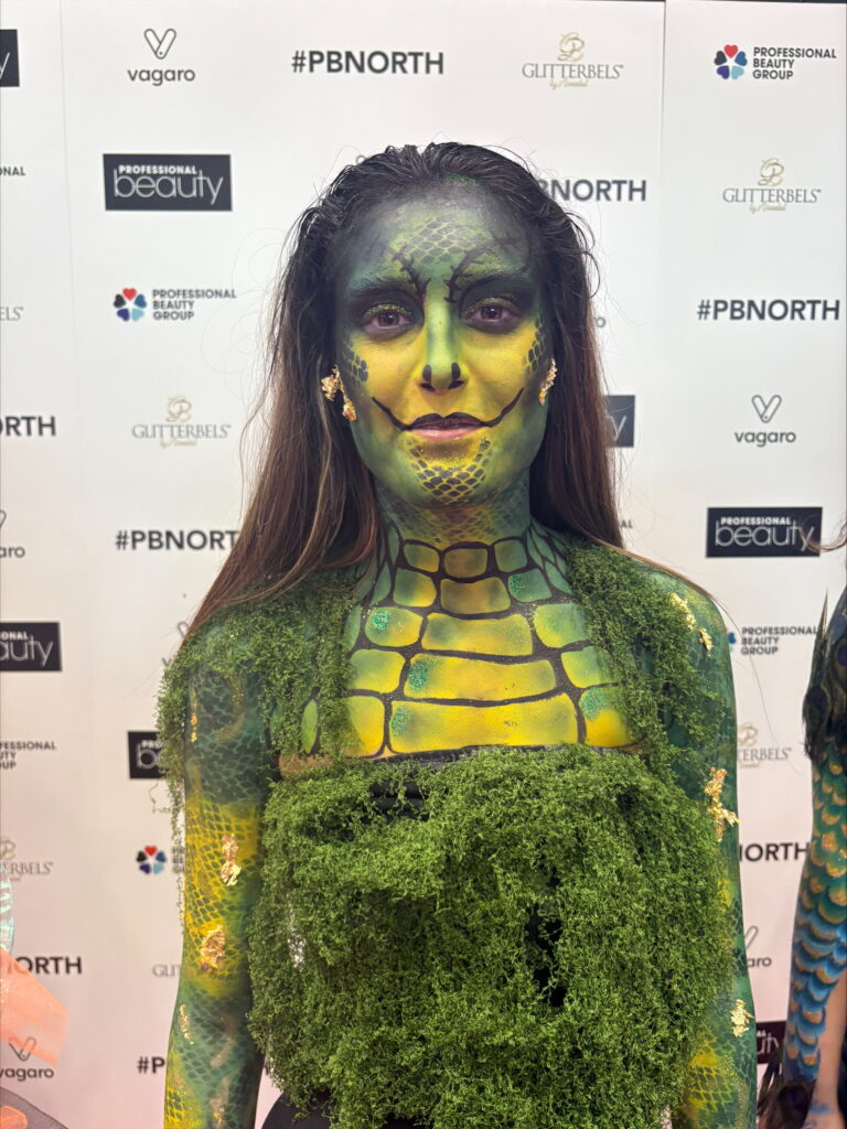 Close-up of a person in green reptilian body paint with moss-like textures and gold facial accents.
