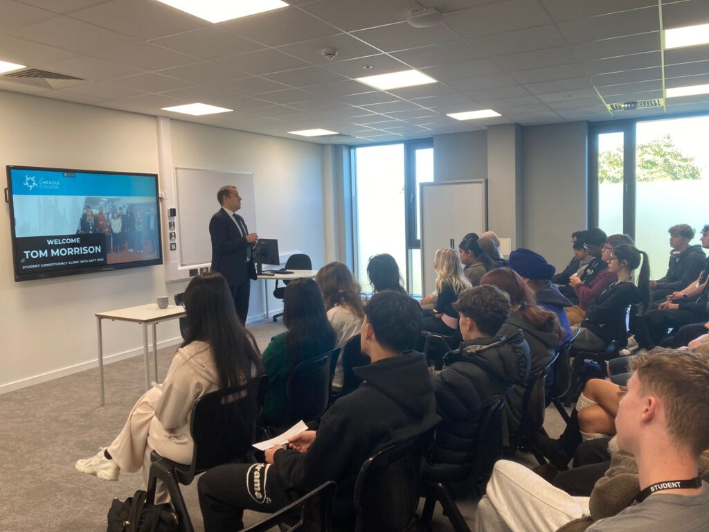 Guest speaker MP Tom Morrison delivering a presentation to college students in a classroom, with a welcome slide displayed on a large screen.