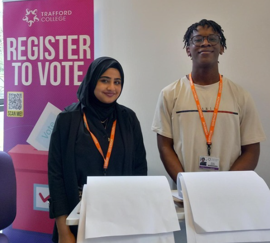 Two students, male and female smiling in front of a register to vote banner
