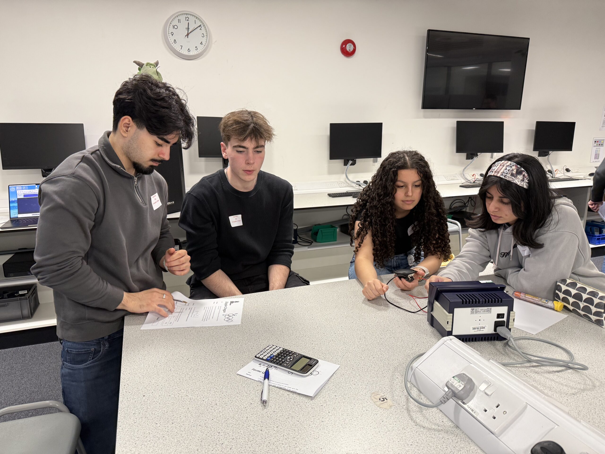 Four individuals are gathered around a gray laboratory table in a classroom setting. The table has a calculator, pen, and paper on it, along with an electronic device connected to wires. The background shows several computer monitors mounted on the wall, a clock, and a power strip on the table. The group appears to be engaged in a hands-on activity involving the equipment.
