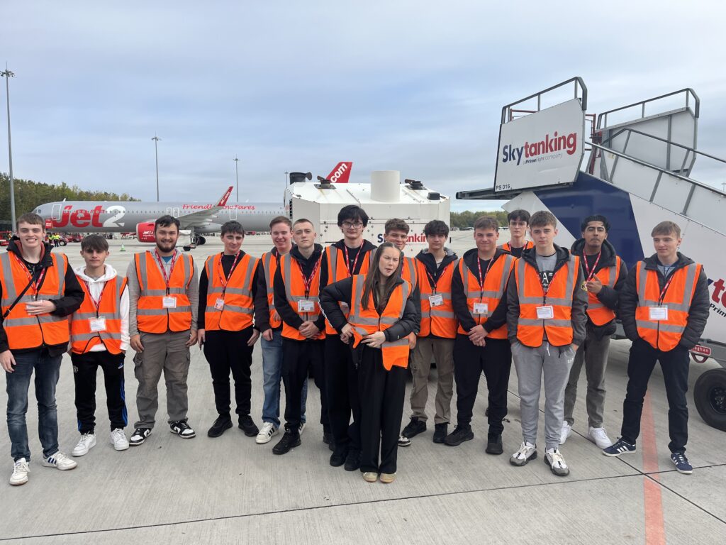 A group of people wearing orange high-visibility vests standing outdoors on an airport apron. Behind them are airport ground service vehicles and a set of aircraft steps marked “Skytanking.” Jet2 aircraft are visible in the background.