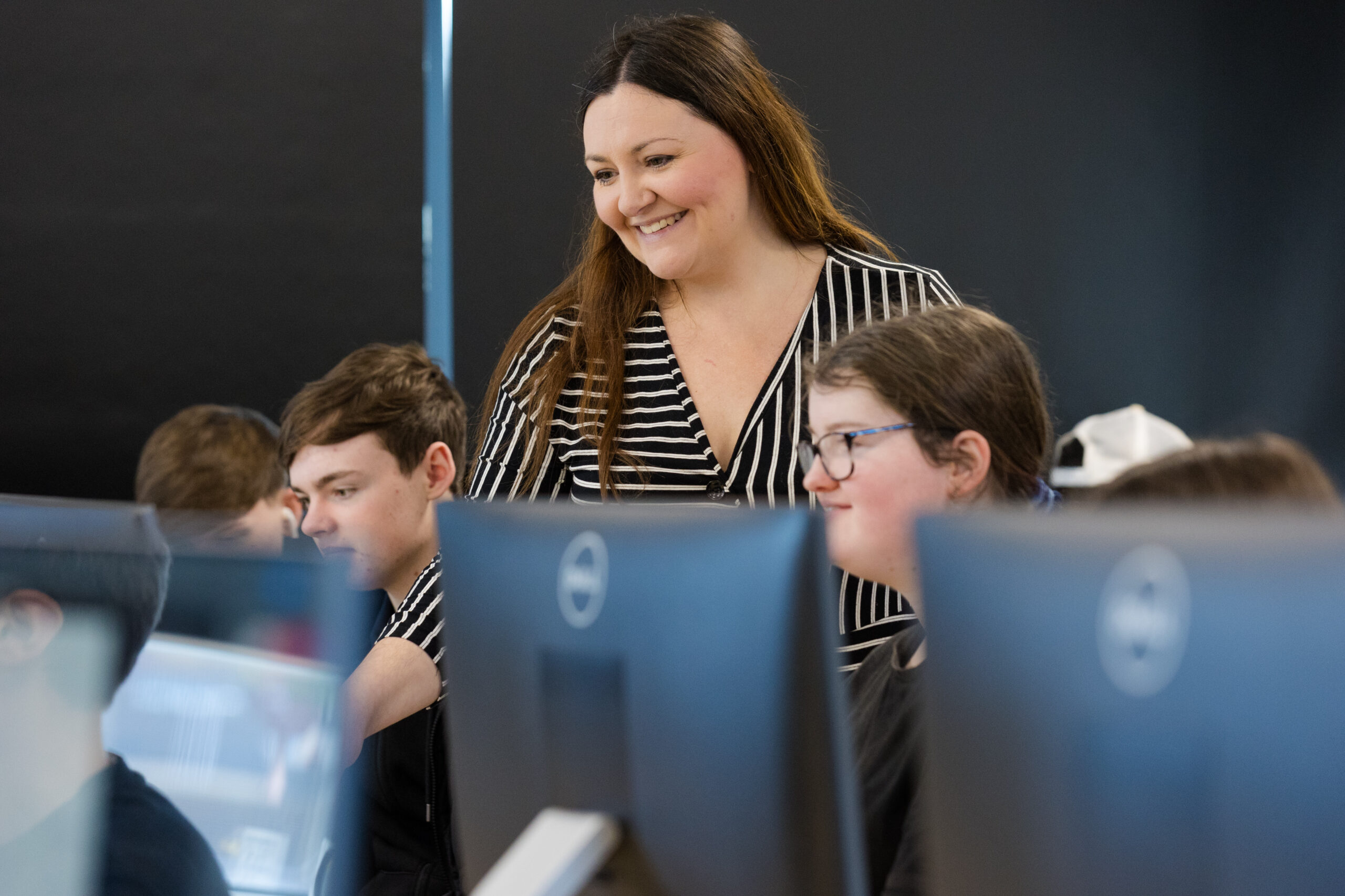 Smiling adult woman stands between two students working at a bank of computer monitors, looking at one o the student's work