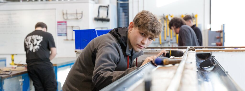 A young man is in a workshop environment, with other students in the background. He is holding a long piece of piping in place and is bent to look down the piping and straight into the camera.