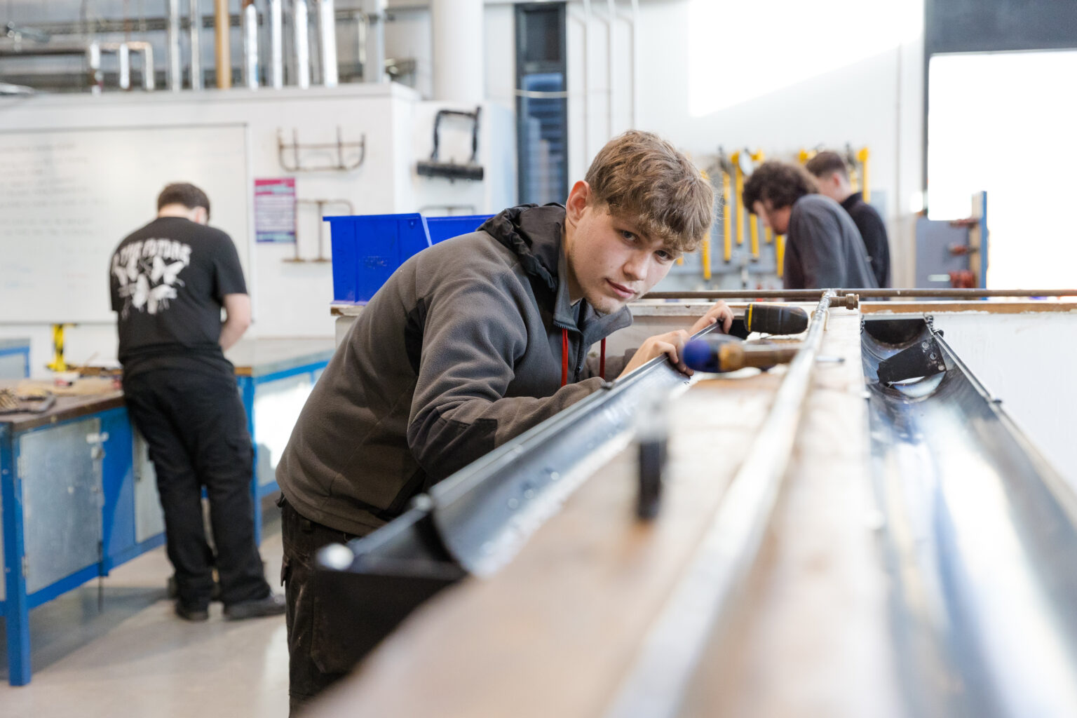 A young man is in a workshop environment, with other students in the background. He is holding a long piece of piping in place and is bent to look down the piping and straight into the camera.
