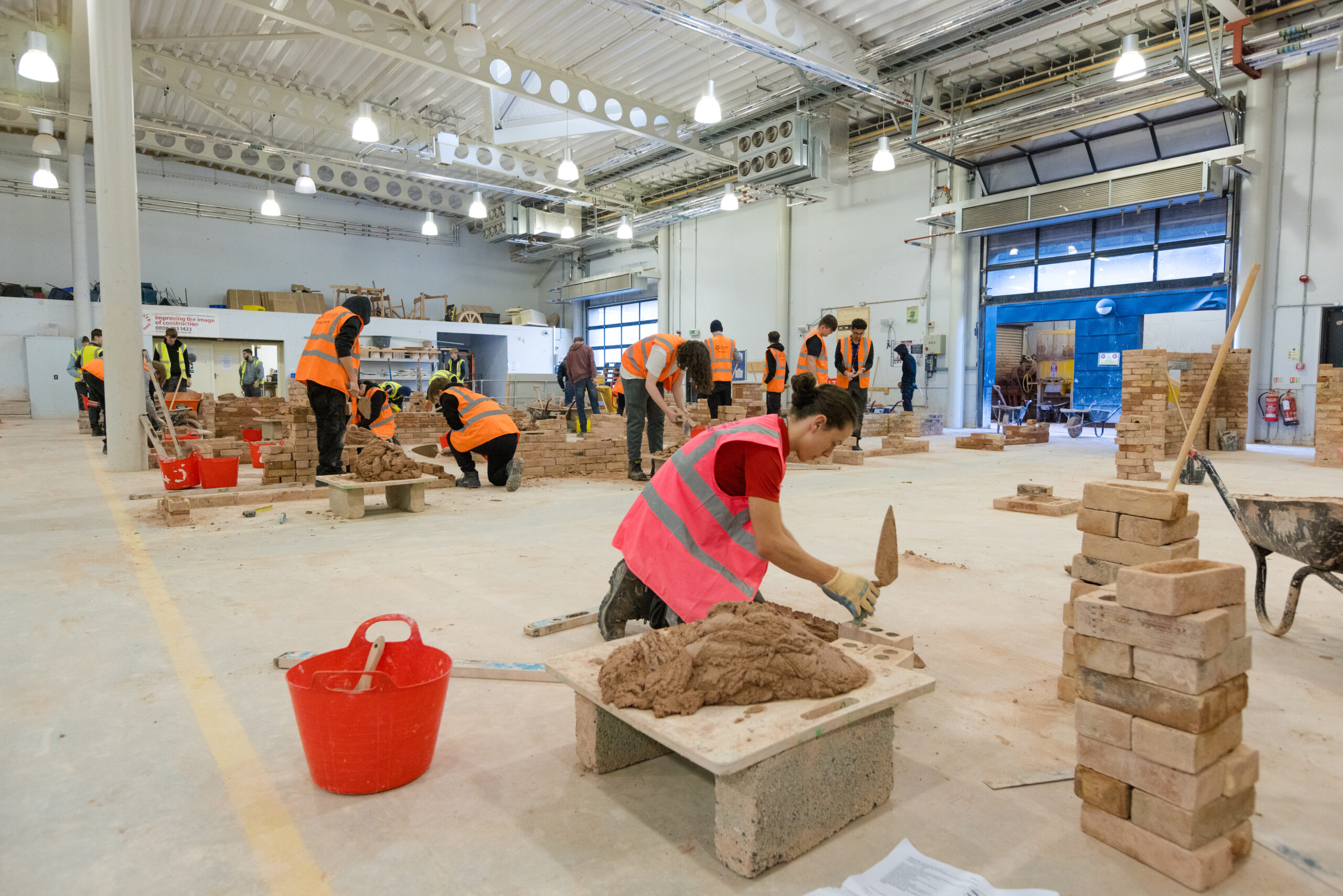 A large brightly lit bricklaying workshop where there are several stations set up with bricks and mortar and equipment for bricklayingg like trowels, water and red buckets. At each station there are students in high vis jackets each at different stages of building a piece of wall with the materials.