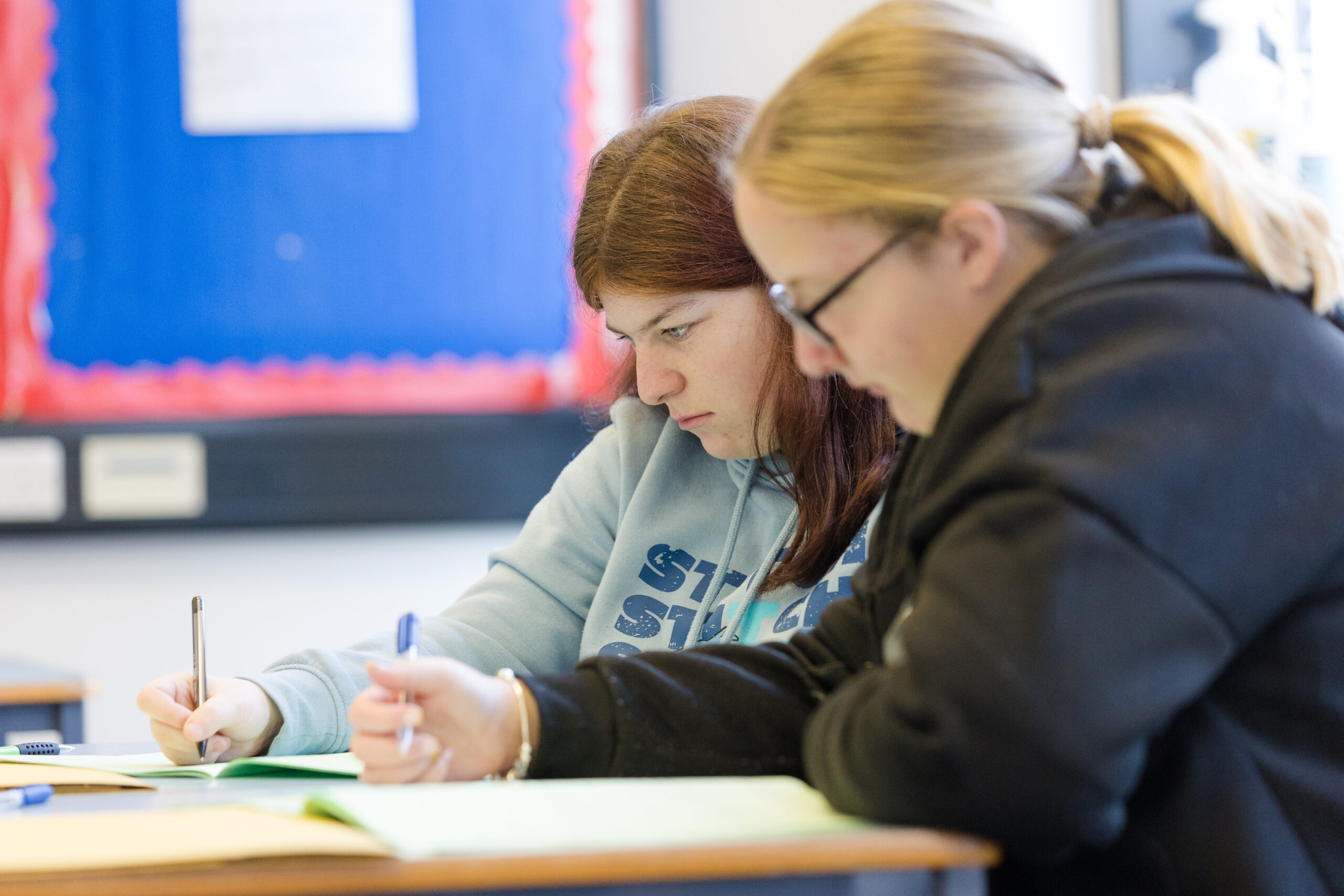 Two female students are sat at a desk in a classroom focused on writing in an exercise book.