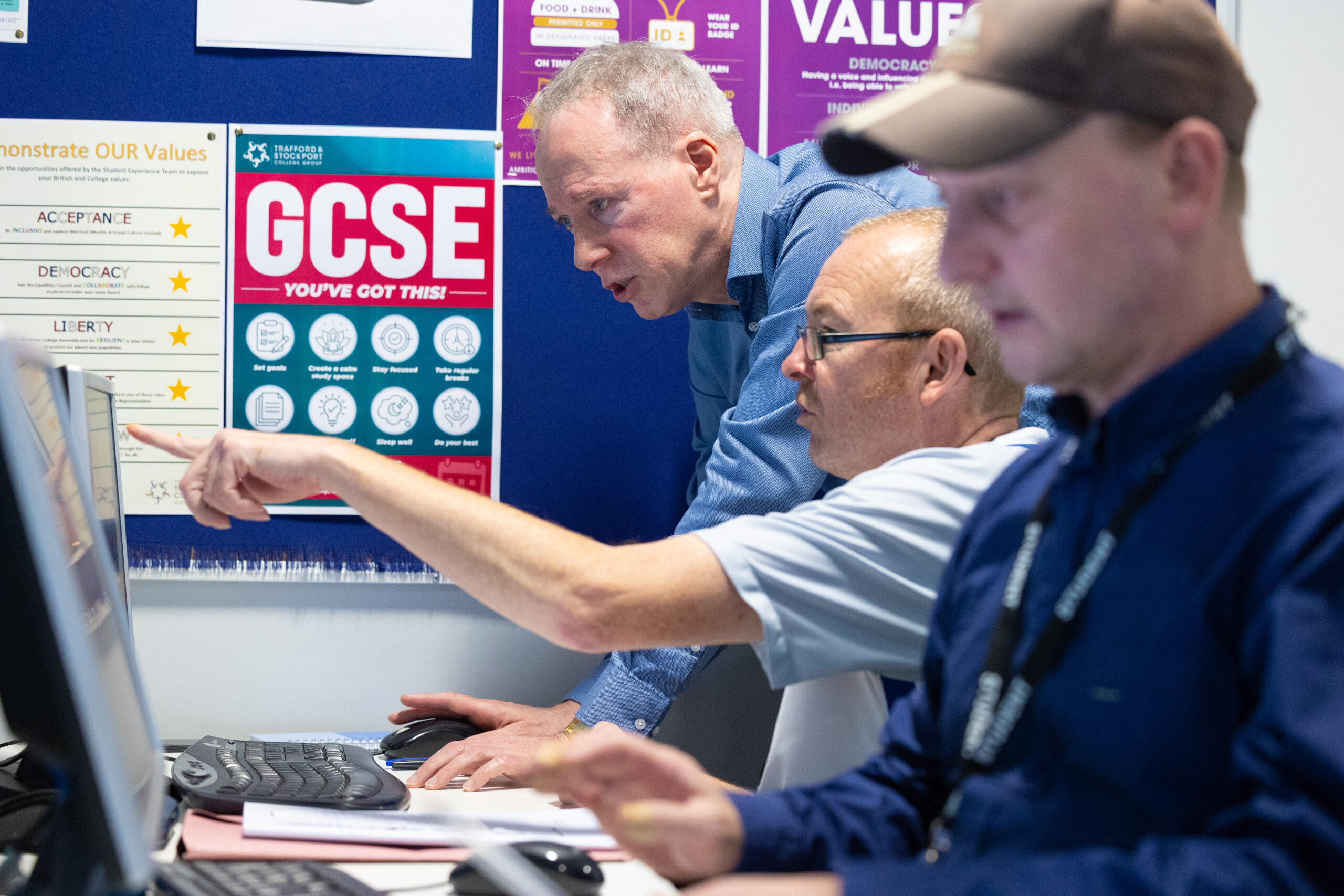 An adult man wearing a blue shirt is stood next to a seated man in front of a computer monitor in a classroom environment. The seated man is pointing to the screen and both men are focused on the screen and discussing. In the foreground, another man is working on another screen.