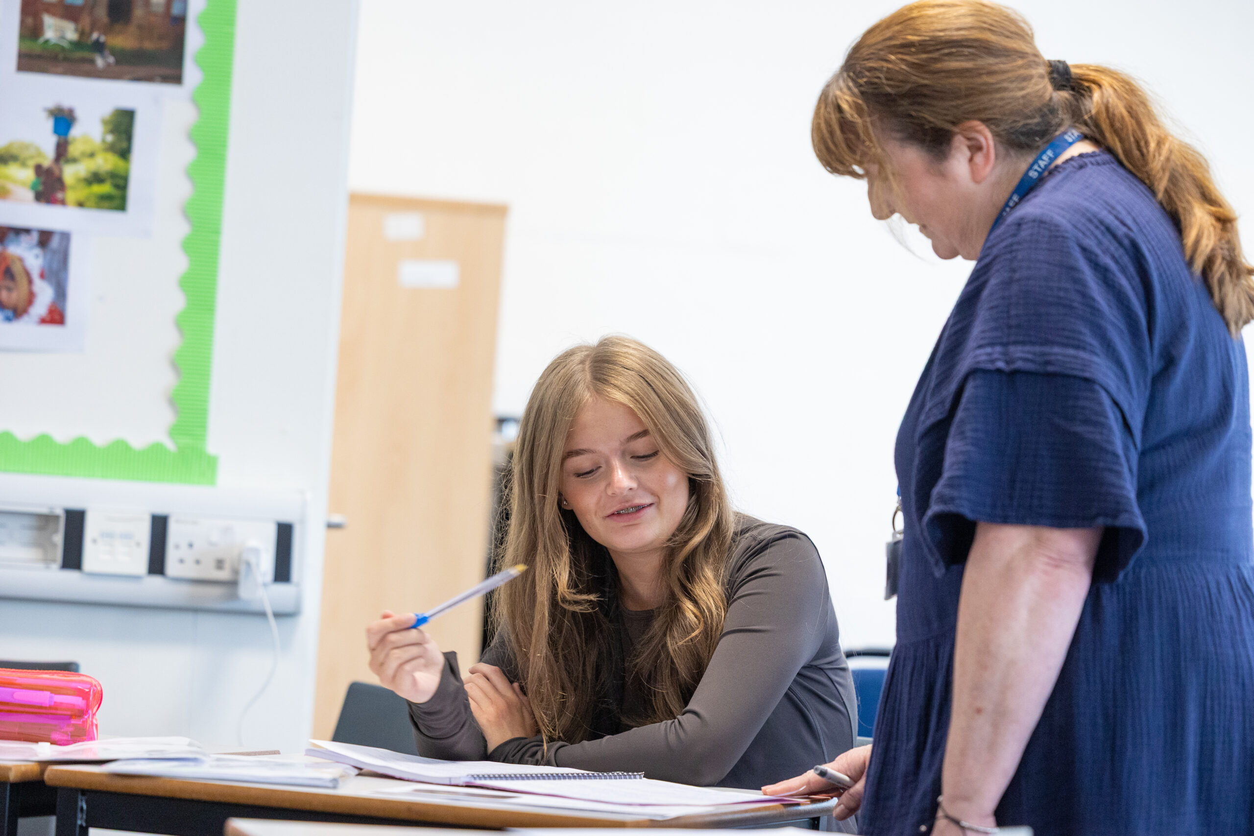 an older woman stands next to a younger female student who is sat at a desk in a classroom. Both are looking down at an exercise book that sits between the two women. The younger woman is talking through her work.