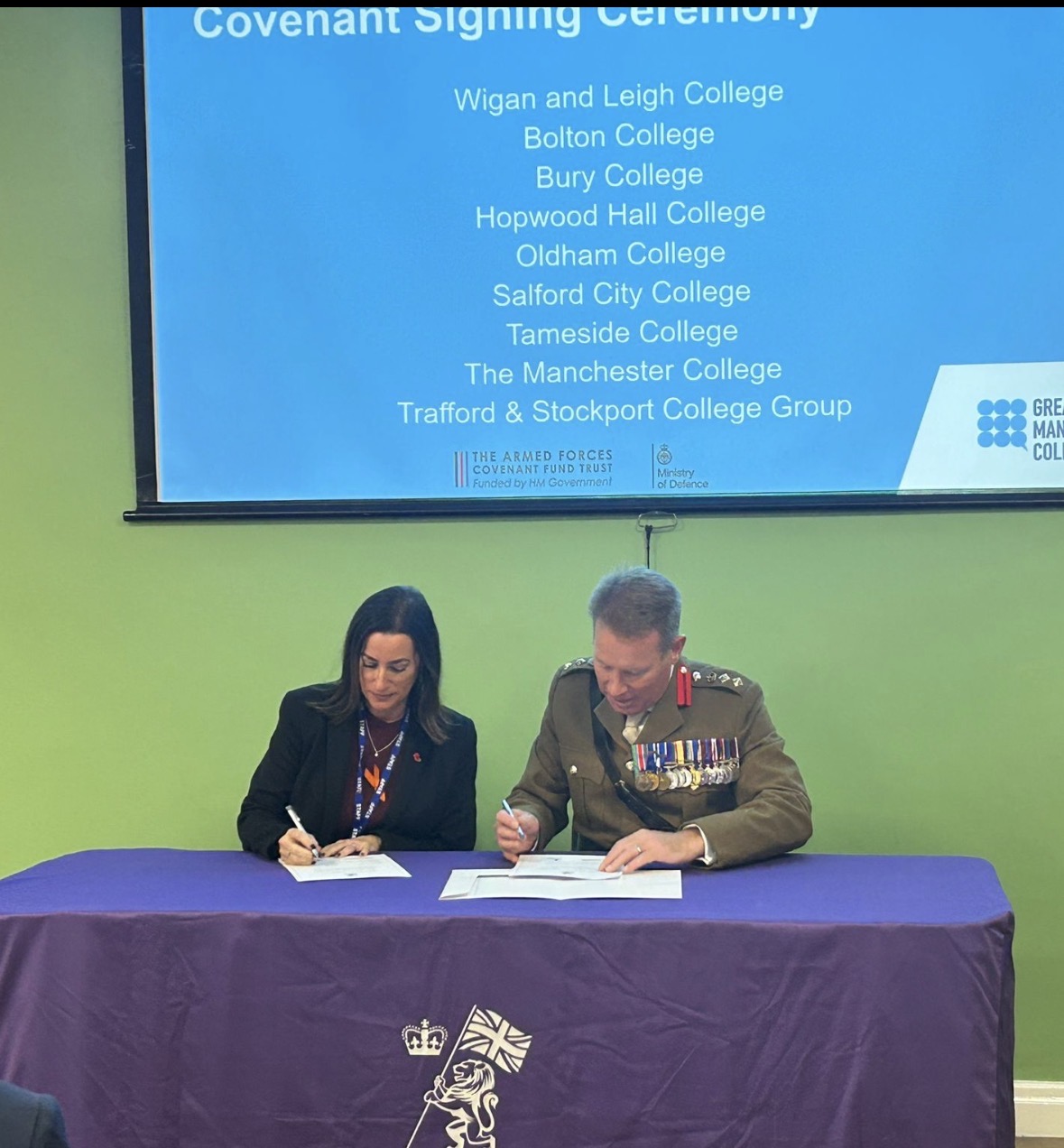 Two individuals signing documents at a table during a Covenant Signing Ceremony, with a presentation screen behind them listing participating colleges.